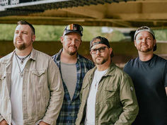 Four men pose under a bridge, wearing casual jackets and caps, with calm expressions in a soft, green outdoor setting.