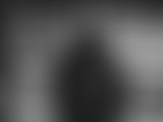 Long-haired person in a black suit stands in front of a rustic wooden wall, looking serious. Black and white image with moody tone.