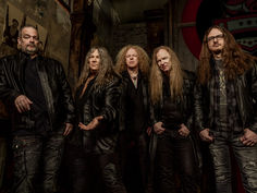 Five long-haired men in black leather jackets pose in a dim, gritty room with a rock-style logo in the background.