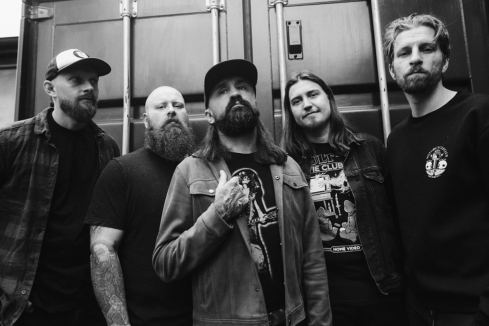 Five men with beards stand closely in front of a container. One wears a shirt with a musician print. The setting is monochrome and moody.