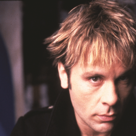 Close-up of a blond man with an intense, serious expression against a dark blurred background.