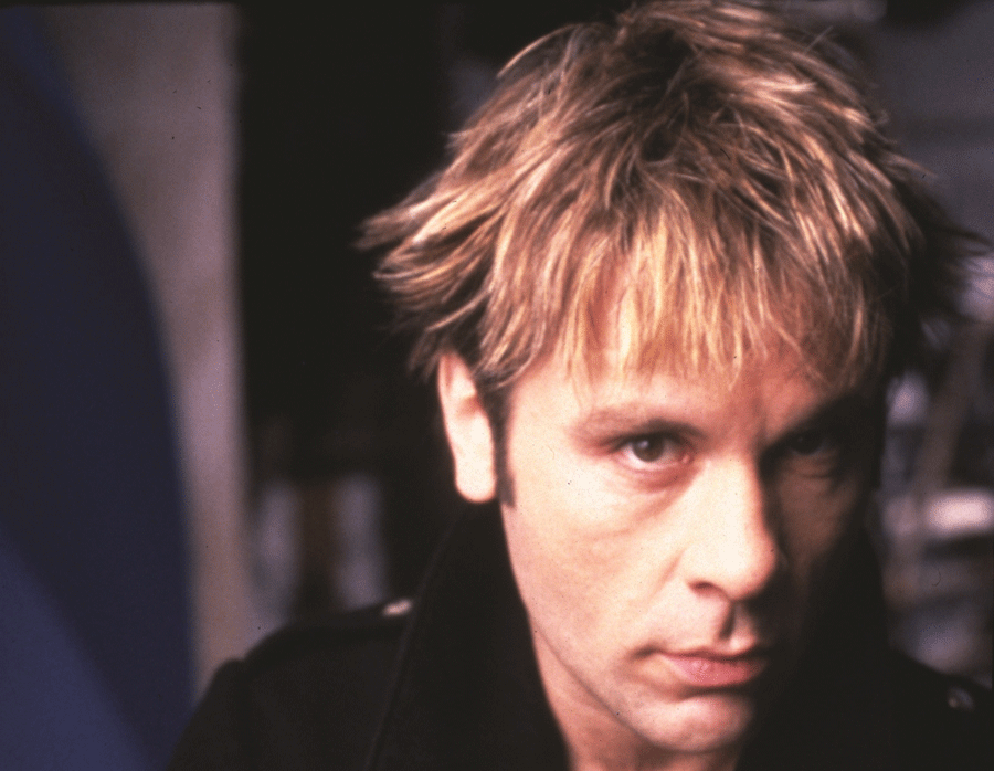 Close-up of a blond man with an intense, serious expression against a dark blurred background.