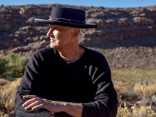 Man in a black hat and sweater gazes to the side, seated in a sunny desert landscape with rocky hills, creating a contemplative mood.