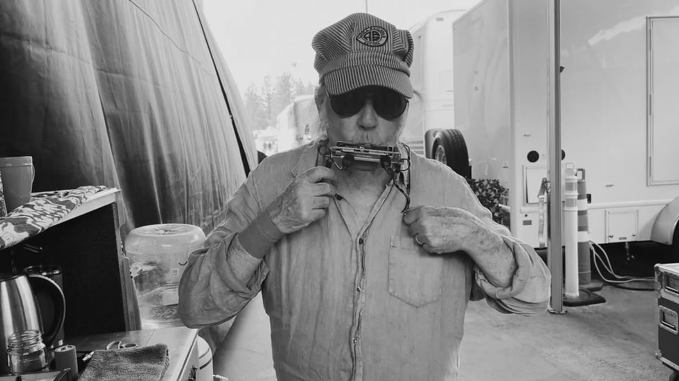 Man with sunglasses and striped cap plays harmonica outdoors. Background has trailers, cones, and a table with mugs and towels. Monochrome.