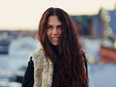 Smiling woman with long dark hair in a beige fur vest, standing on a rooftop with blurred city lights at sunset.