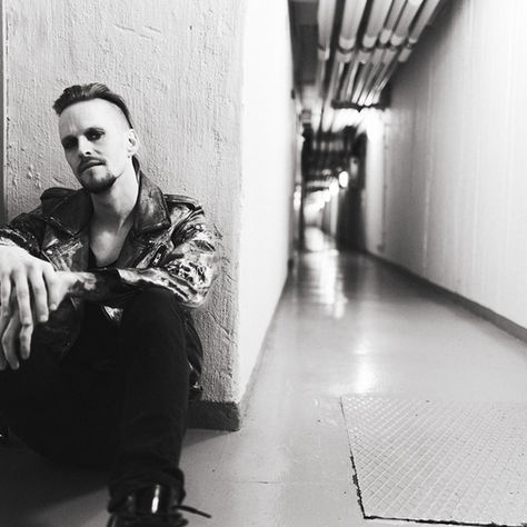 Man in leather jacket sits on floor in a dim hallway. Blank expression. Pipes line the ceiling, creating a tunnel effect. Black and white.