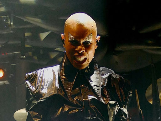 Bald person in shiny black attire with dramatic eye makeup performs on stage, set against dark background with warm lighting. Mood is intense.
