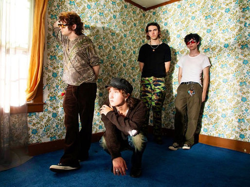 Four people in a room with floral wallpaper and blue carpet. One crouches, others stand. Casual attire, relaxed mood, sunlight through curtains.