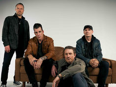 Four men pose around a brown couch in a studio, wearing dark jackets, with serious expressions against a plain white background.