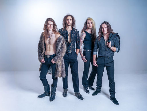 Four men with long hair pose confidently in stylish outfits against a plain blue backdrop. One wears a fur coat, others have tattoos.