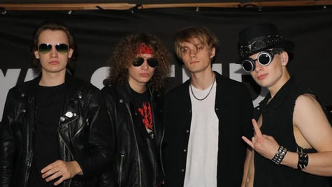 Four men in leather attire pose in front of a black backdrop. Two wear sunglasses, one has a red headband, another wears a top hat. Rock vibe.