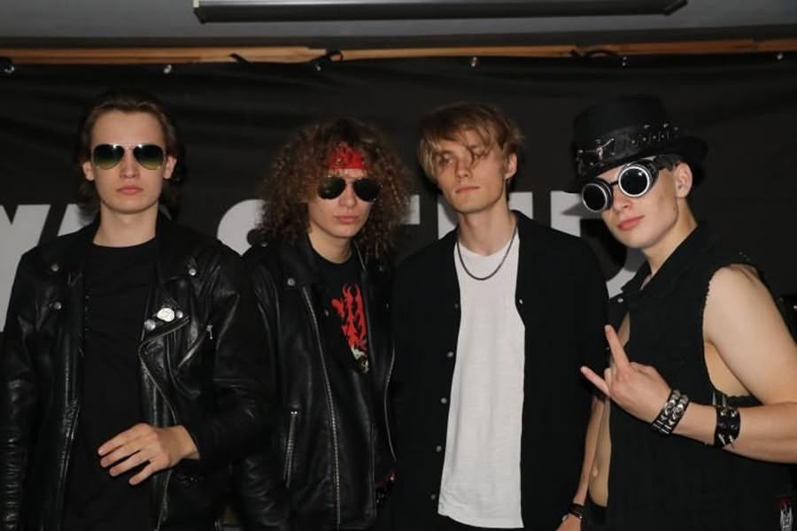 Four men in leather attire pose in front of a black backdrop. Two wear sunglasses, one has a red headband, another wears a top hat. Rock vibe.