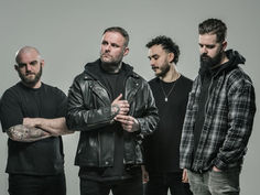 Four men in dark clothing stand against a gray background. One wears a leather jacket, others checkered or plain shirts. Serious expressions.