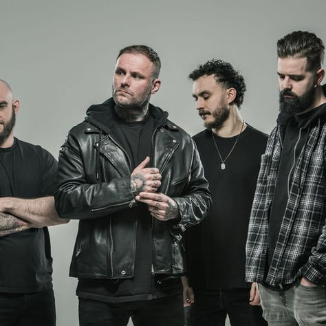 Four men stand against a gray background. They wear dark clothing, including a leather jacket and plaid shirt, and display calm expressions.