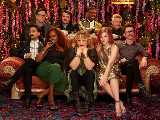 Eight adults pose on a pink velvet couch amid pink flower garlands and warm lights, wearing glam outfits, in a festive studio scene.