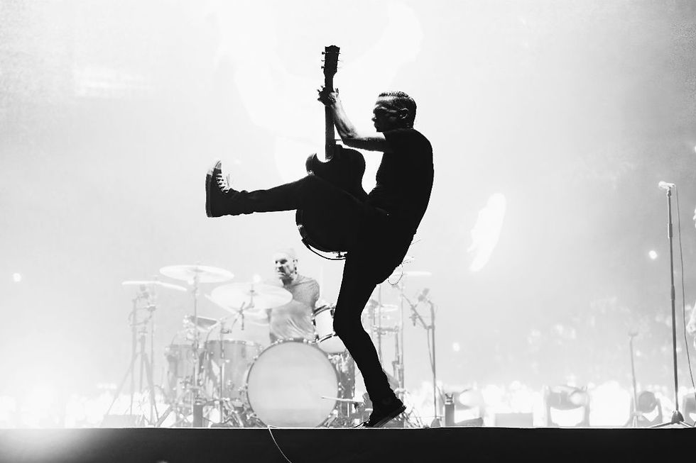 Black-and-white concert scene: guitarist leaps midair with one leg outstretched, drummer behind, bright stage lights.