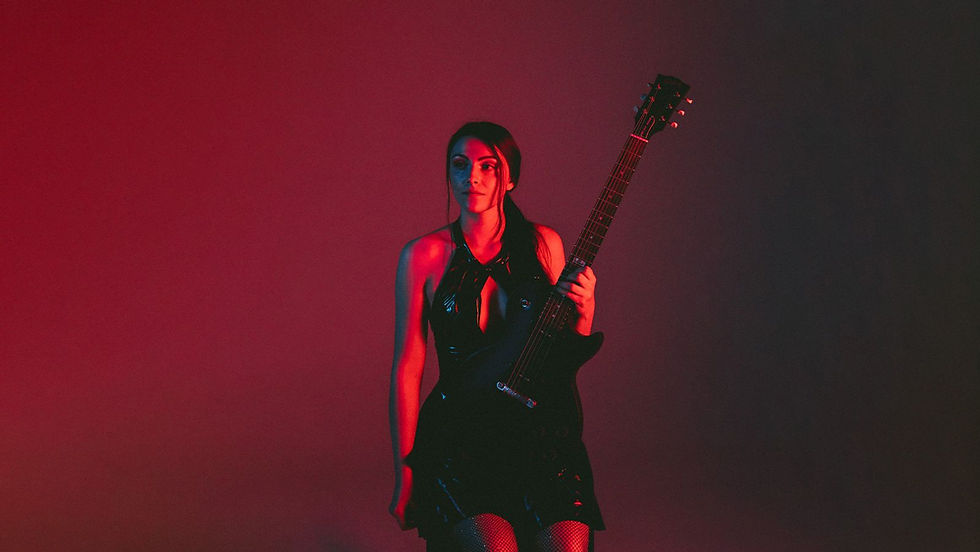 Woman in a black dress holding an electric guitar against a red-pink studio backdrop, lit with moody neon light.