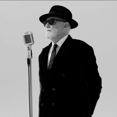 Black-and-white portrait of an older man in a hat and sunglasses standing at a vintage microphone against a plain backdrop.