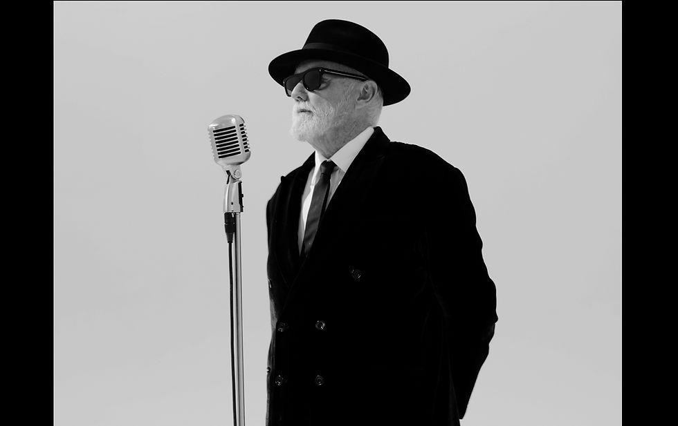 Black-and-white portrait of an older man in a hat and sunglasses standing at a vintage microphone against a plain backdrop.