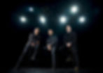 Three men in black leather jackets sit on stools under bright stage lights against a dark background, exuding a serious, confident vibe.