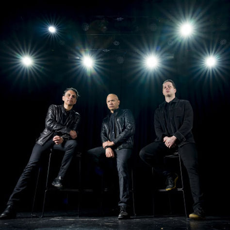 Three men in black leather jackets sit on stools under bright stage lights against a dark background, exuding a serious, confident vibe.