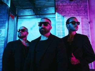 Three men in black suits and sunglasses stand confidently under blue and purple neon lights. A "No Parking" sign is visible.