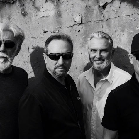 Four men in sunglasses stand against a weathered wall. One wears a hat with "Deus" text. The mood is serious and monochrome.