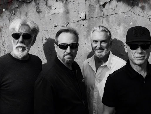 Four men in sunglasses stand against a weathered wall. One wears a hat with "Deus" text. The mood is serious and monochrome.