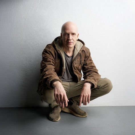 Bald man squats in casual brown attire against a plain gray wall, casting soft shadows. Neutral expression, relaxed mood.