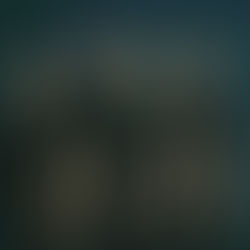 Four men in black shirts stand together against a dark teal background, displaying serious expressions. Their arms hang relaxed by their sides.