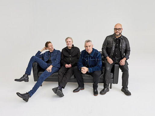 Four men in dark clothes sit on a black couch in a white studio, posing casually.