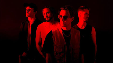 Four men stand in dramatic red lighting against a dark background, exuding a mysterious and intense mood.