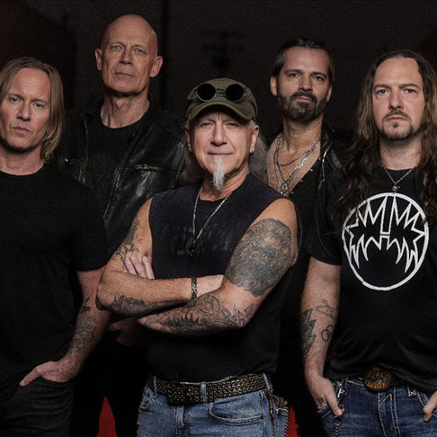 Five rock musicians pose in black shirts against a dark red backdrop, arms crossed and serious, with tattoos and a band logo tee.