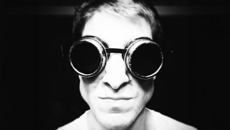 Person wearing black goggles in a close-up, monochrome image. The background is dark, and the mood is mysterious and intense.