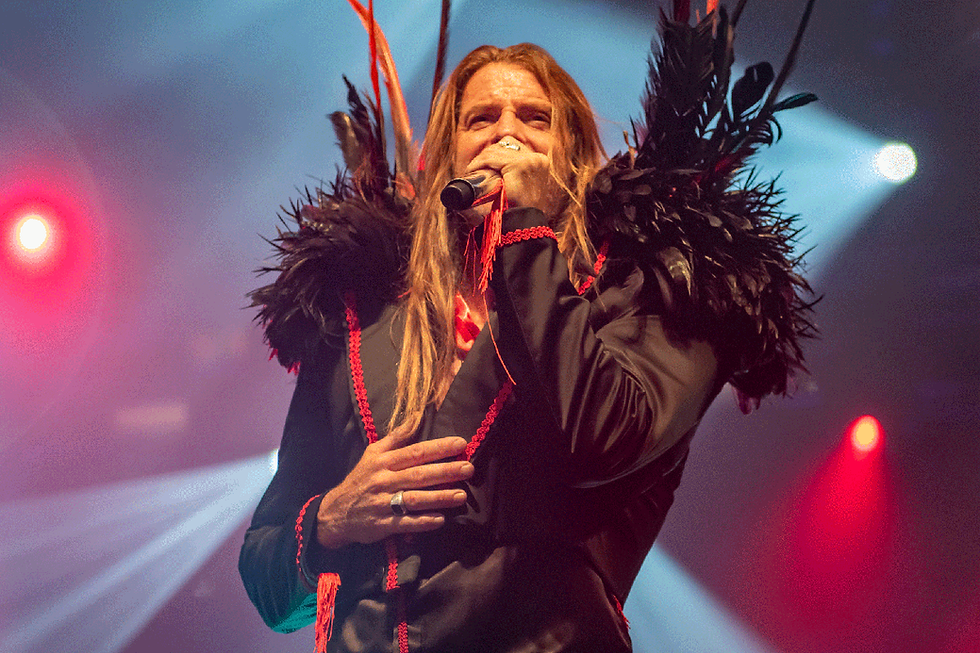 Performer with long hair sings passionately on stage, wearing a black feathered outfit with red accents. Bright stage lights in the background.