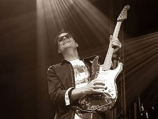 Guitarist in sunglasses playing energetically on stage under dramatic spotlights, exuding joy. Sepia tone adds vintage feel.