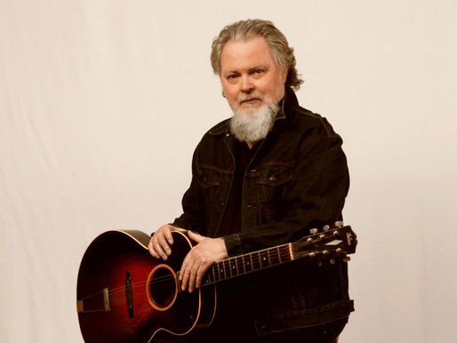 Bearded man in a black jacket holds a vintage acoustic guitar against a plain beige backdrop, exuding a calm and relaxed mood.