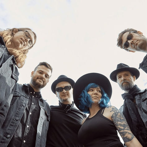 Band members in black outfits pose outdoors under a cloudy sky. One has blue hair and a tattoo, others wear sunglasses and hats. Mood is edgy.