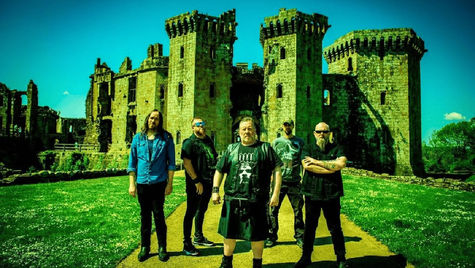 Five men stand on a path with a medieval castle in the background. The sky is clear blue, giving the scene a dramatic and bold mood.