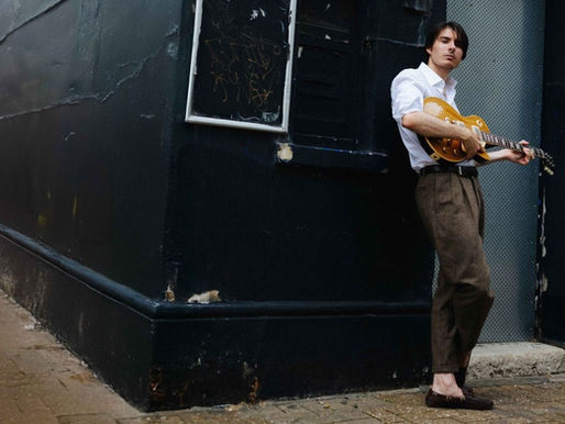 Young man in a white shirt leans against a dark wall, playing a guitar. He stands on a brick sidewalk, exuding a relaxed, contemplative mood.