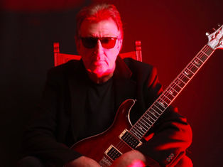 Man in sunglasses with a guitar, seated against a black and red-lit background. He wears black, exuding a confident and moody vibe.