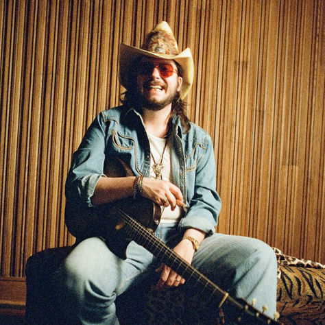 Smiling man in a cowboy hat and sunglasses sits with a guitar in a wood-paneled room, relaxed and playful.
