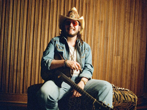 Smiling man in a cowboy hat and sunglasses sits with a guitar in a wood-paneled room, relaxed and playful.