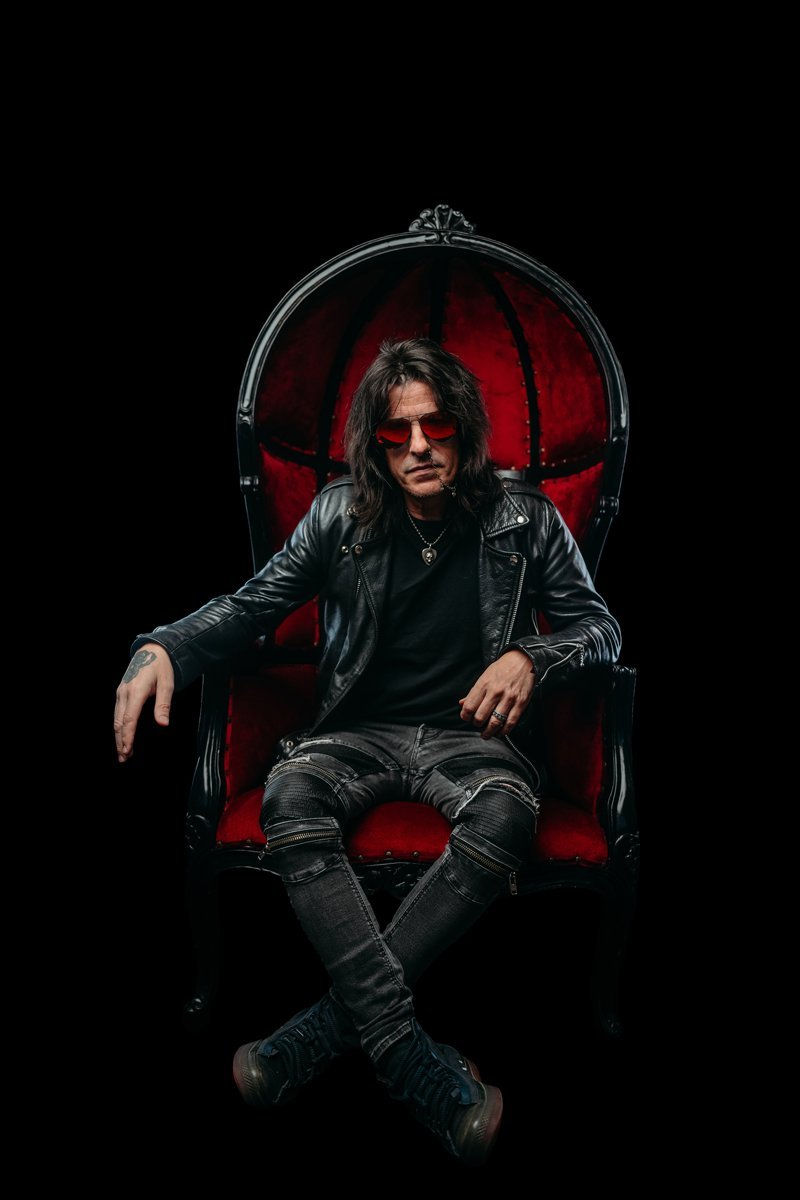 Man in red sunglasses and leather jacket sits confidently on a red velvet chair against a black background, exuding a rock vibe.