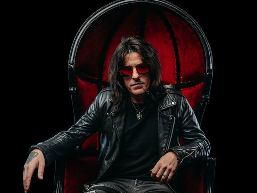 Man in red sunglasses and leather jacket sits confidently on a red velvet chair against a black background, exuding a rock vibe.