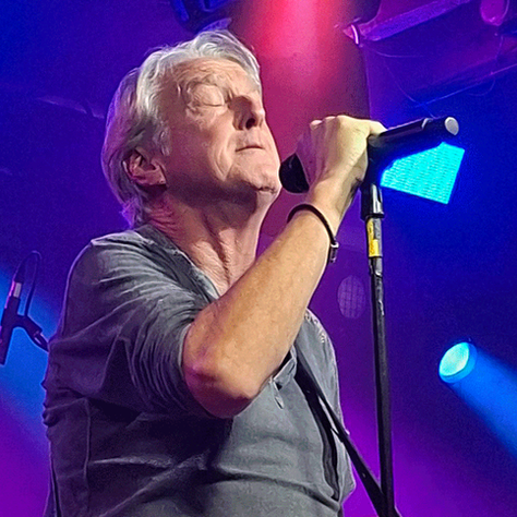 Singer passionately performing on stage, holding a microphone. Colorful spotlights in blue and pink create a vibrant concert atmosphere.