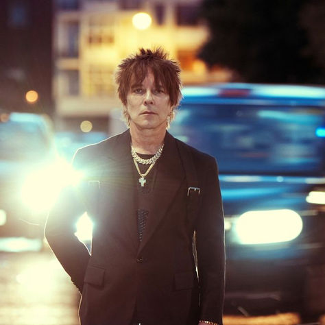 Man in a black suit stands on a city street at night, illuminated by bright headlights. Background shows blurred vehicles and buildings.