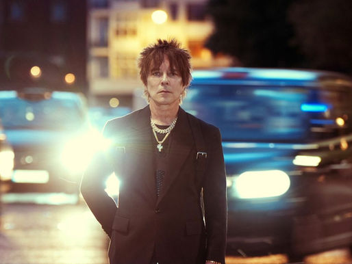 Man in a black suit stands on a city street at night, illuminated by bright headlights. Background shows blurred vehicles and buildings.