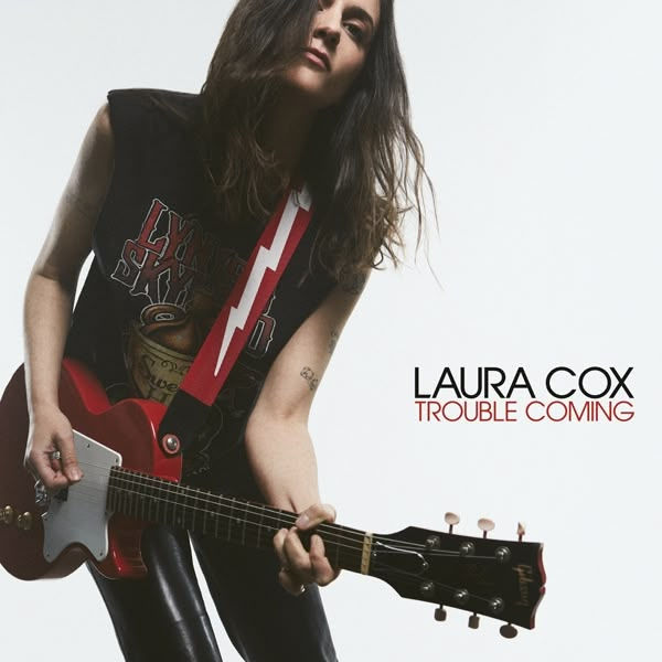 Woman playing guitar with red strap, wearing a black band tee. Text reads: "LAURA COX TROUBLE COMING." White background. Rock vibe.