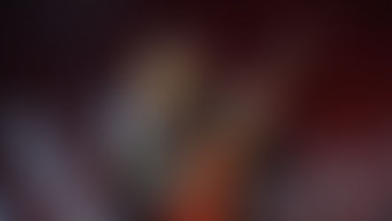 Musician passionately plays a red guitar on stage, eyes closed, wearing a dark polka-dot shirt. Background has large, blurred text.